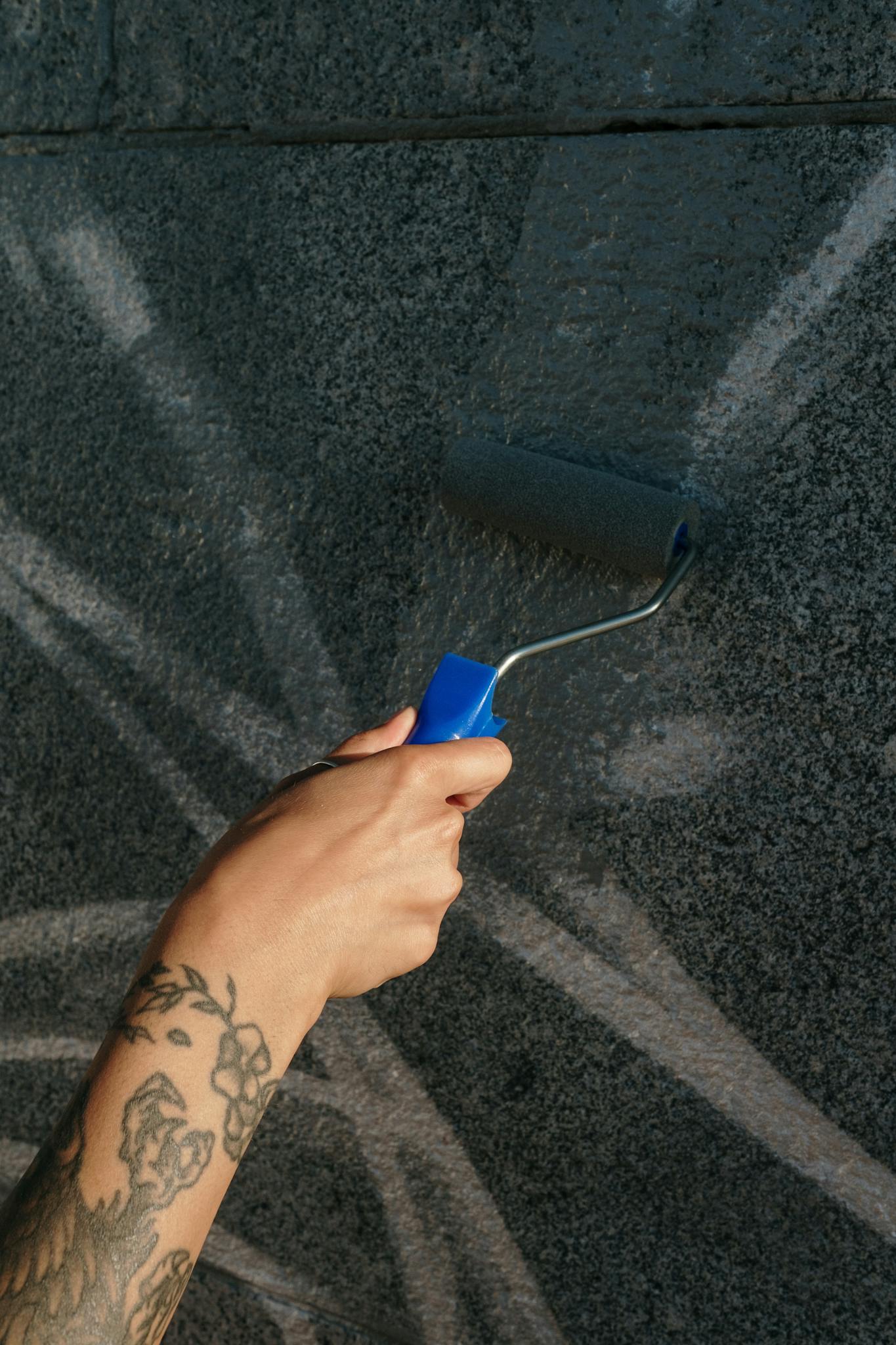 A tattooed hand using a paint roller to cover graffiti on a cement wall.
